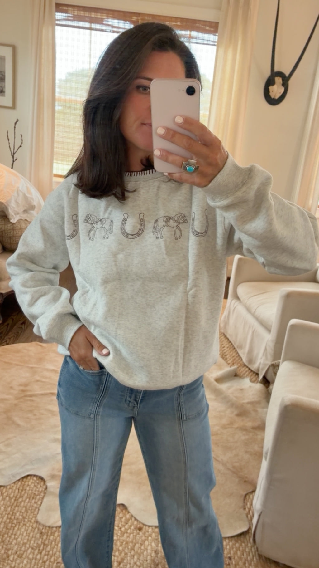 A woman takes a mirror selfie wearing a light gray heathered sweatshirt with a rhinestone-embellished chest graphic and blue jeans in a cozy, sunlit living room.