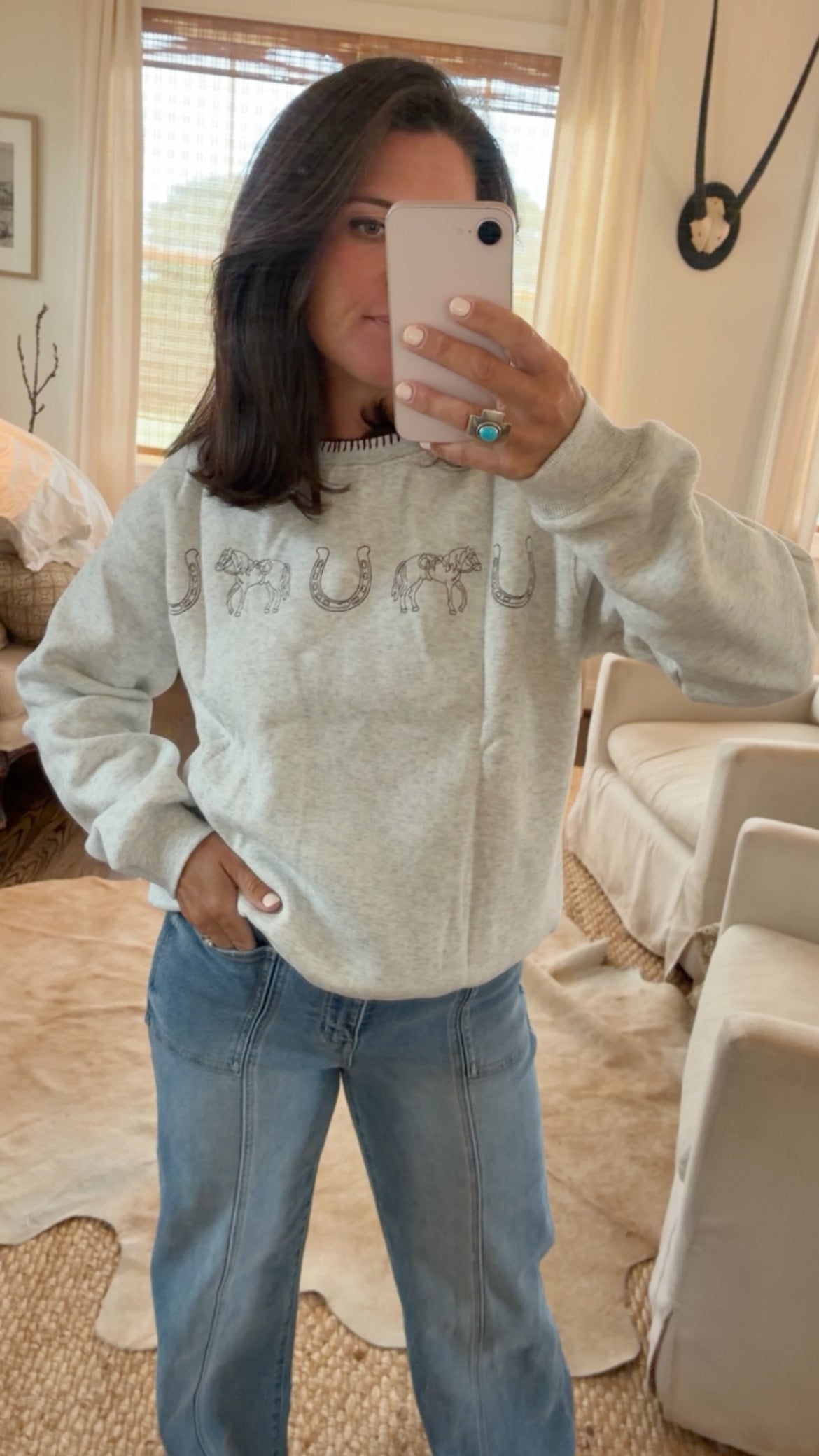 A woman takes a mirror selfie wearing a light gray heathered sweatshirt with a rhinestone-embellished chest graphic and blue jeans in a cozy, sunlit living room.