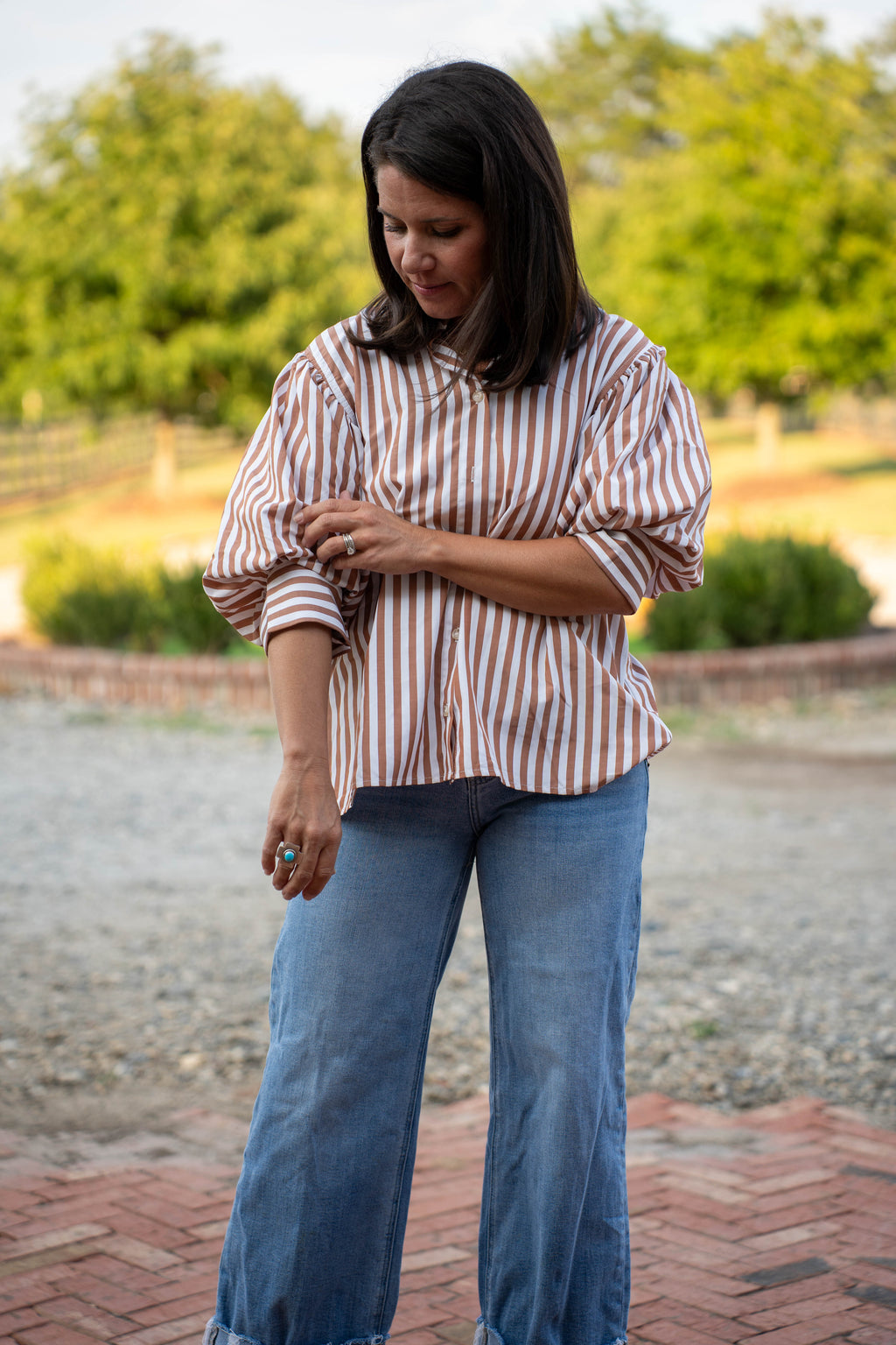 A woman wearing a brown and white vertically striped button-up shirt with voluminous puff sleeves and blue jeans, standing outdoors on a brick and gravel path with trees in the background.
