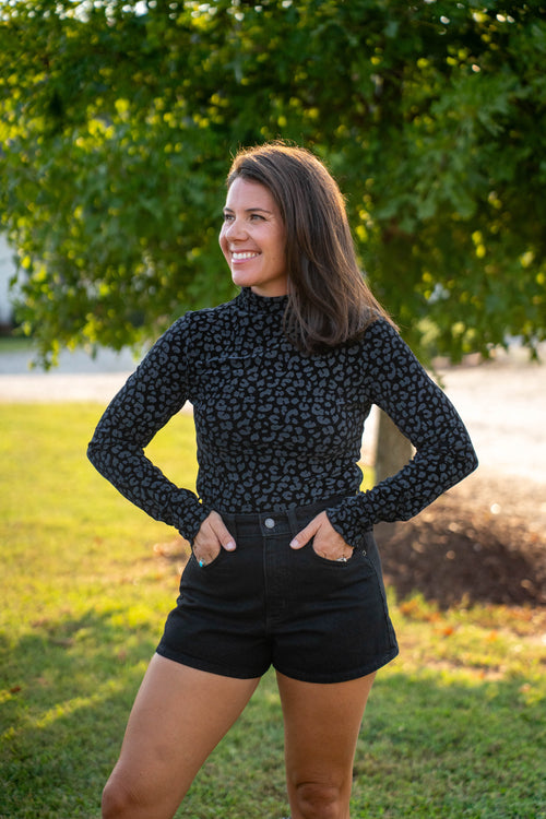 A woman wearing a black long-sleeve mock neck top with a small gray leopard print pattern and black denim shorts, standing outdoors on a grassy lawn with a tree in the background.