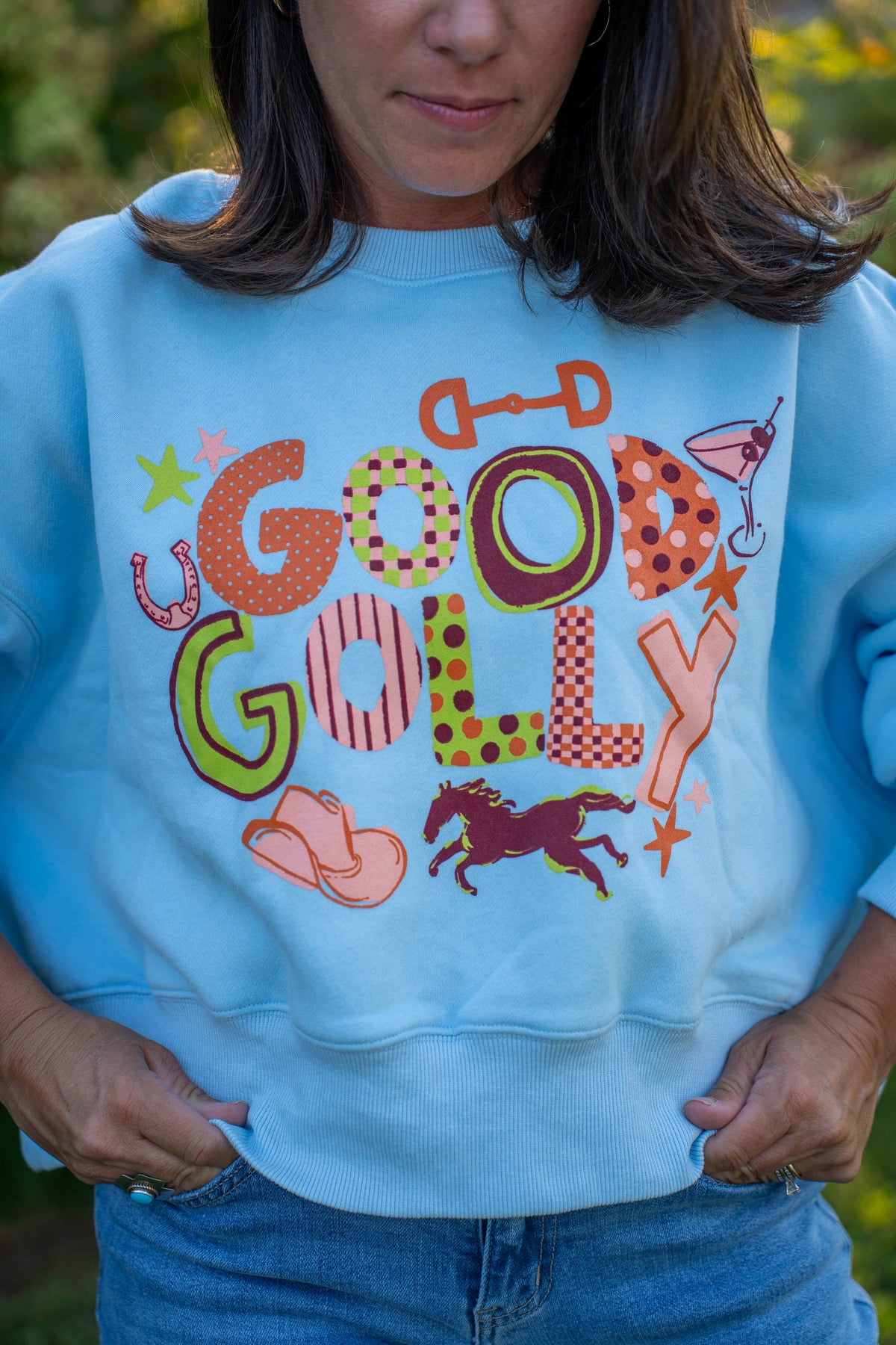 A woman wearing a light blue crewneck sweatshirt with a colorful 'GOOD GOLLY' western-themed graphic print, paired with blue jeans, set against a blurred outdoor background.