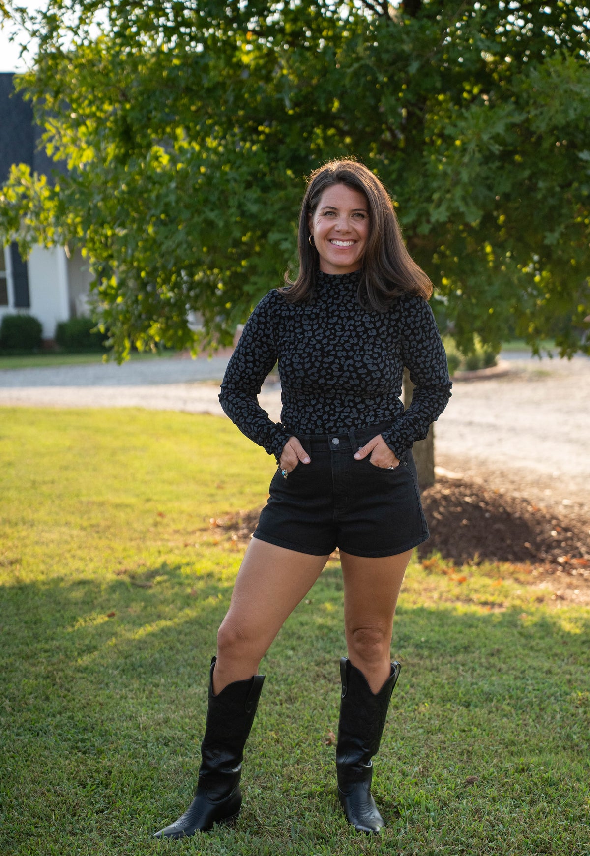 A woman stands outdoors on a grassy lawn in front of a large tree, wearing a black long-sleeve top with a small repeating pattern, black shorts, and black cowboy boots.
