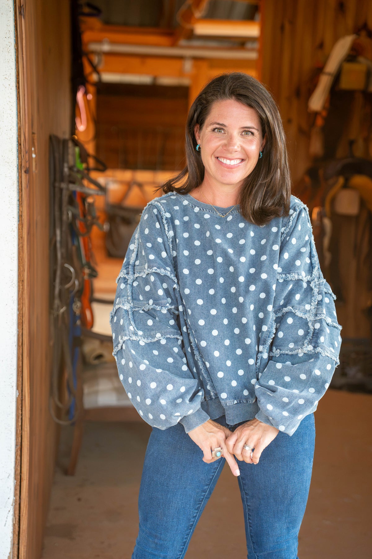 A smiling woman with shoulder-length brown hair stands in a rustic workshop doorway wearing a dusty blue long-sleeve top with white polka dots and tiered ruffle details on the sleeves, paired with blue jeans.