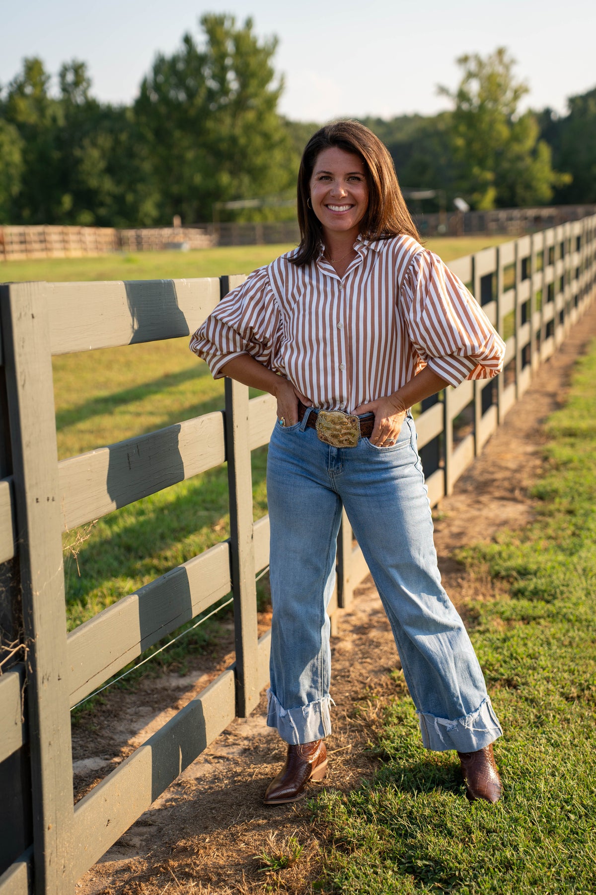 A woman stands outdoors in a grassy field beside a wooden fence, wearing a red and white vertically striped long-sleeve blouse with puffed shoulders, light blue wide-leg jeans with rolled cuffs, and brown boots.