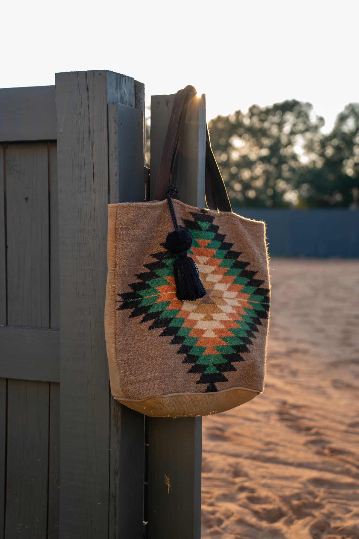 A woven tote bag with a Southwestern geometric diamond pattern in green, orange, and black on a natural beige jute background, hanging from a dark gray wooden fence post in an outdoor setting with sand and trees in the background.