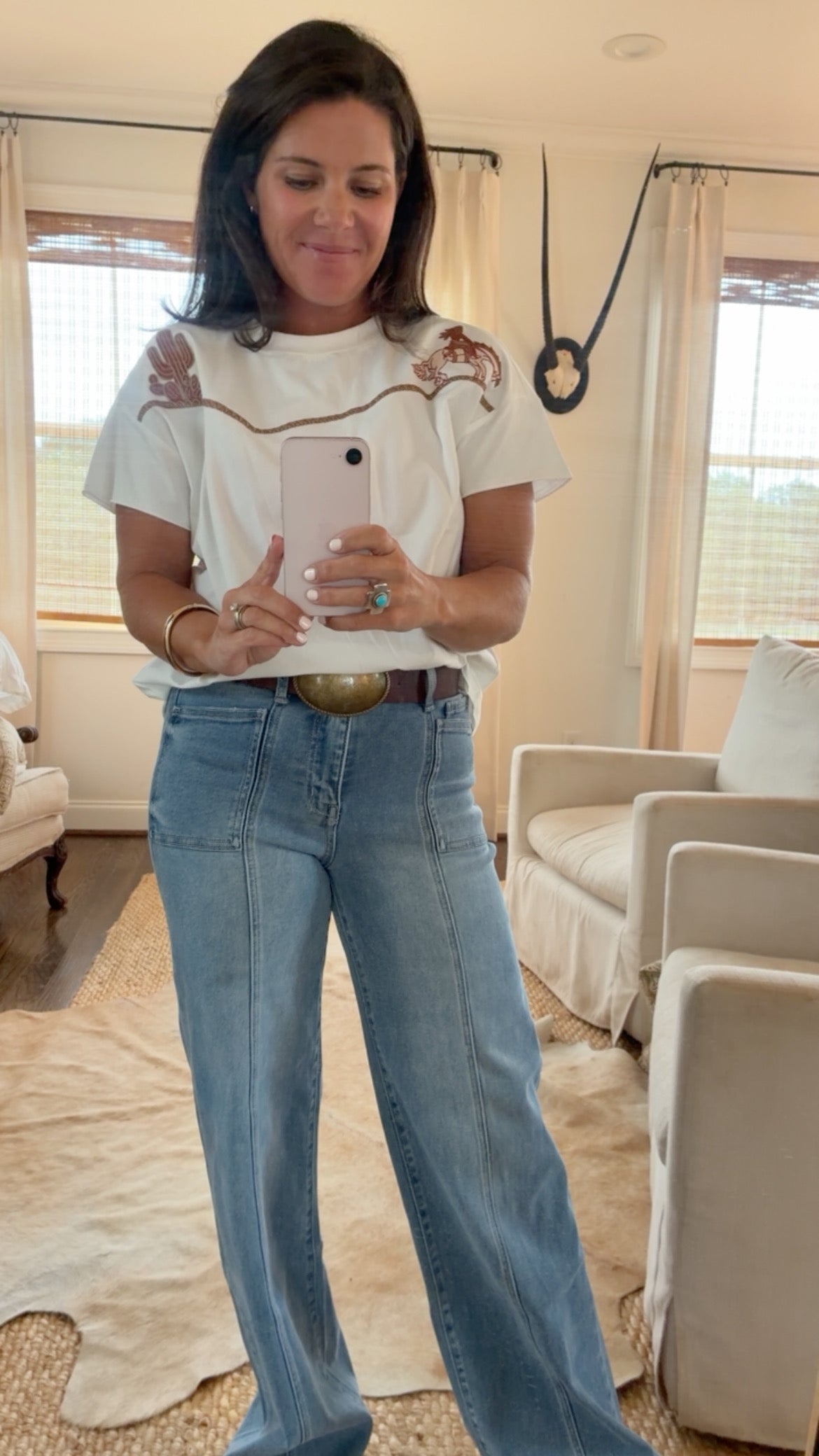 A woman taking a mirror selfie in a living room, wearing a white t-shirt with brown embroidered details on the shoulders and blue wide-leg jeans.