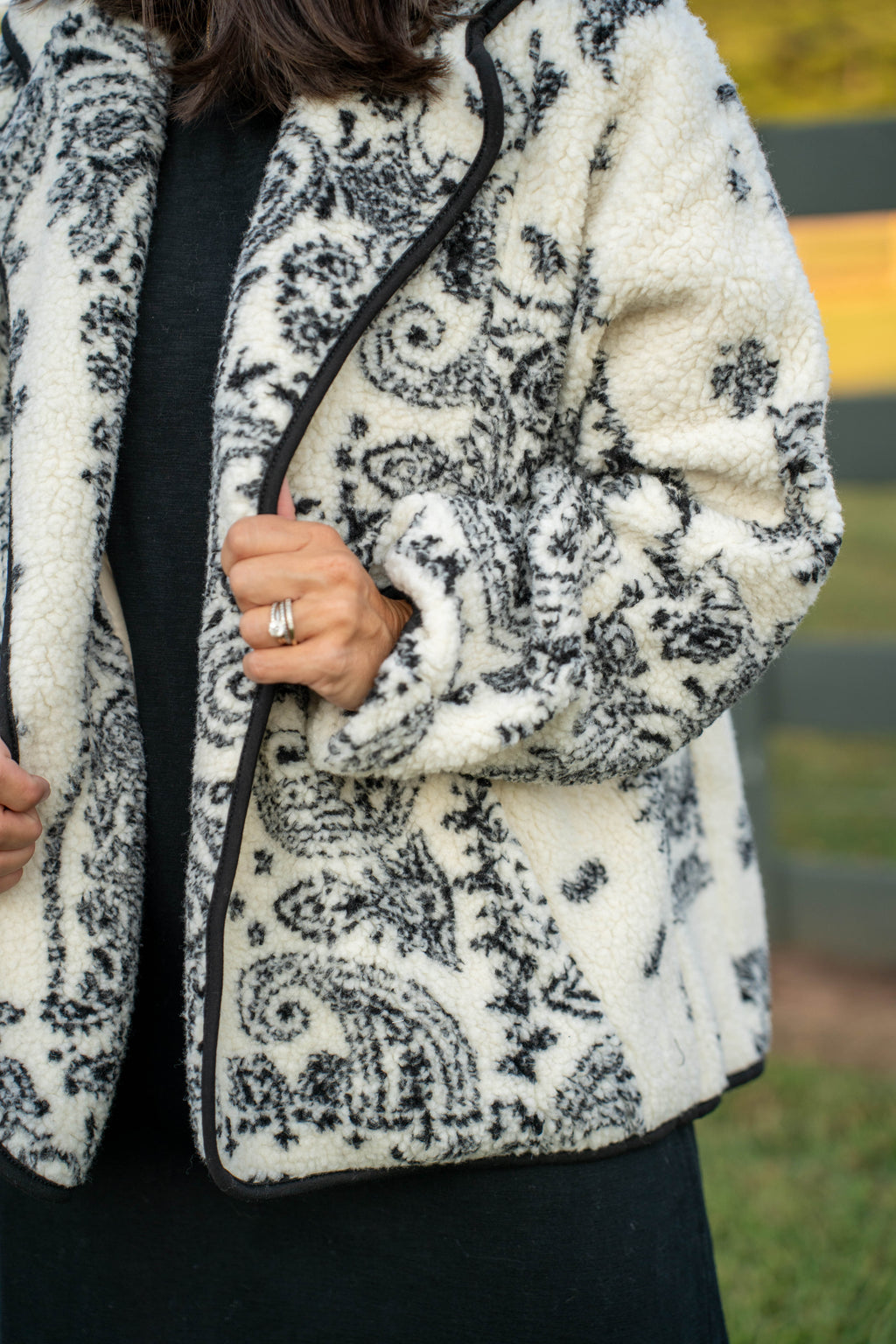 A close-up of a person wearing a cream sherpa fleece jacket with a black paisley pattern and black trim, paired with a black top and skirt. The background is a softly blurred outdoor setting.