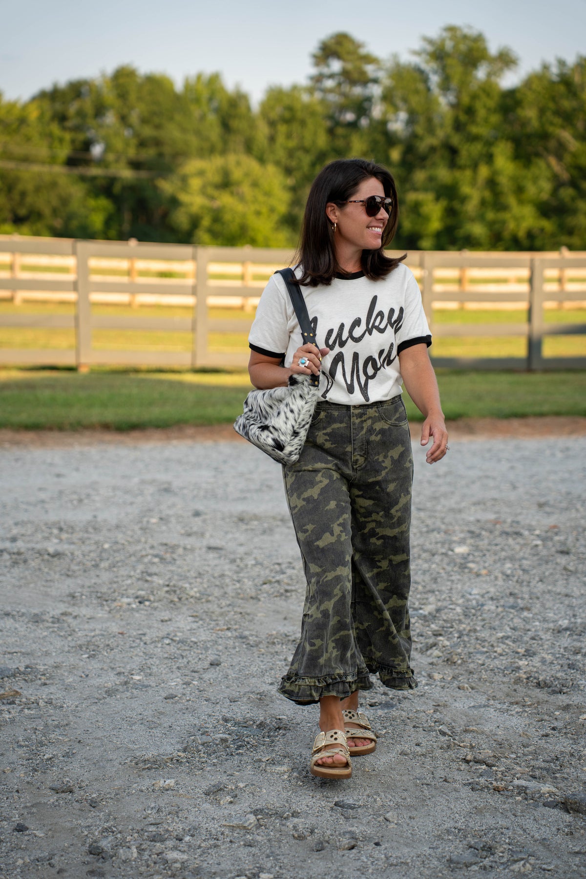 A woman walking outdoors on a gravel path wearing a white t-shirt with black script reading 'lucky mare', camouflage wide-leg pants, and carrying a black and white patterned shoulder bag.