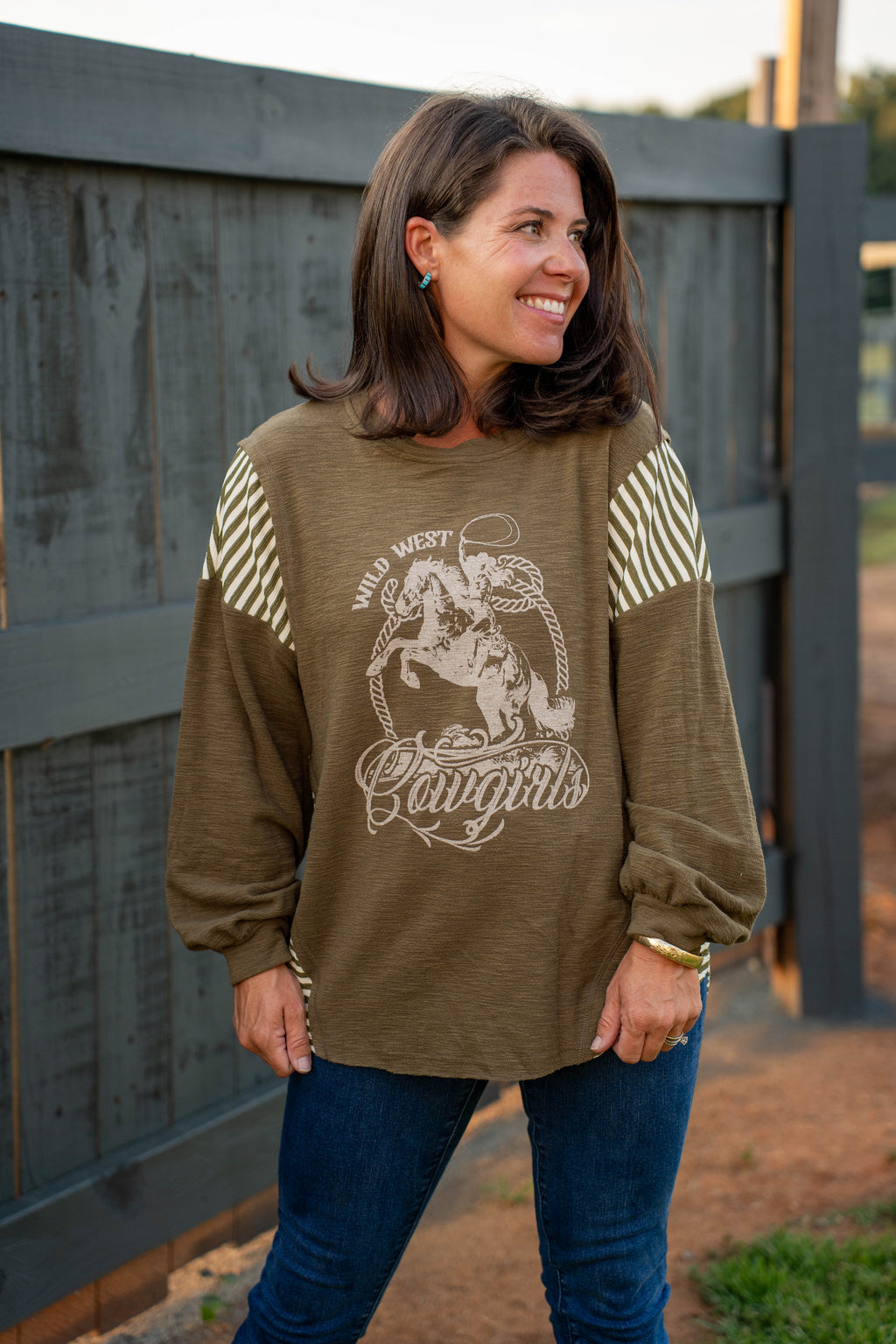 A woman wearing an olive brown long-sleeve graphic sweatshirt with cream and white striped sleeves, paired with blue jeans, standing in front of a dark wooden fence.