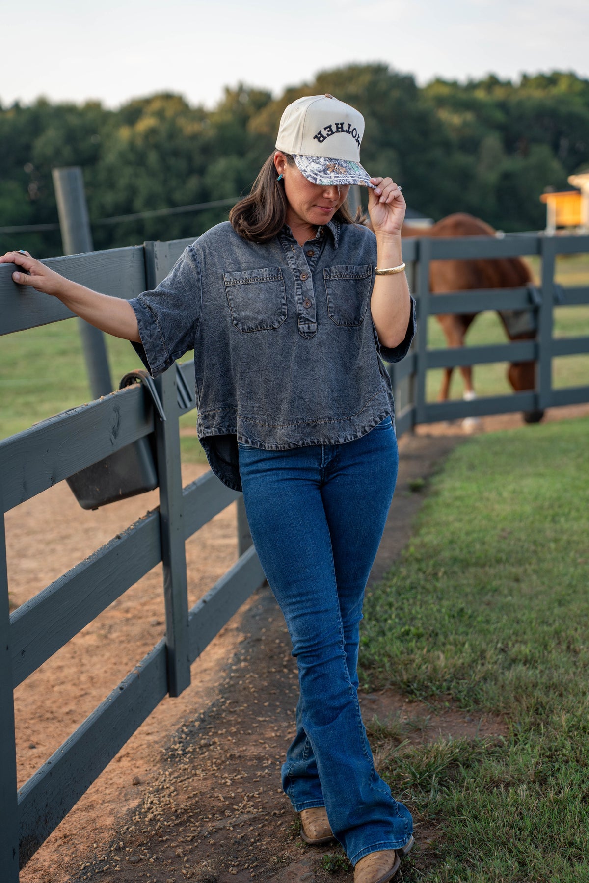 A woman posing outdoors in a rustic paddock next to a wooden fence, wearing a gray washed denim shirt, blue flare jeans, and a beige baseball cap with 'RESILIENCE' printed on it.