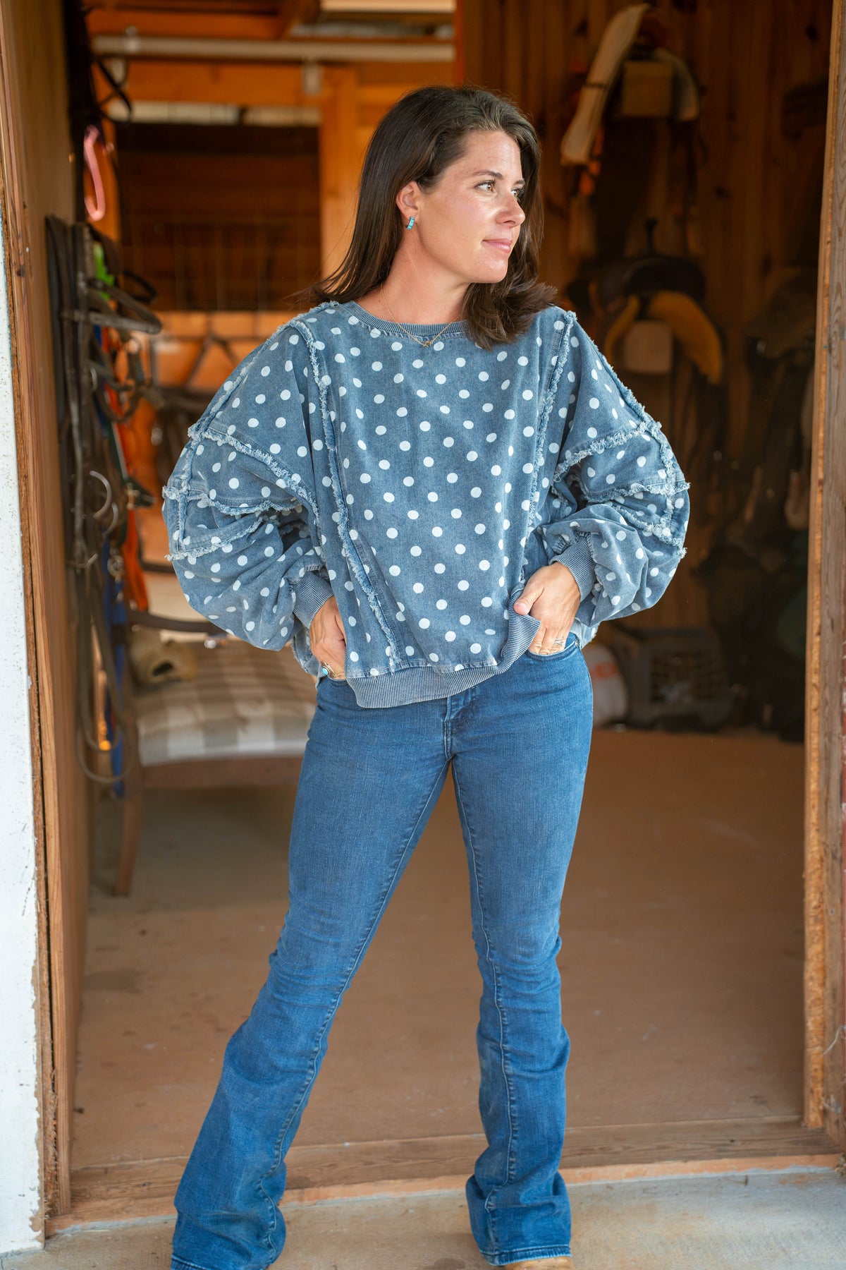 A woman wearing a dusty blue long-sleeve sweatshirt with white polka dots and blue jeans, standing in a rustic wooden doorway.