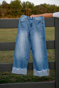 Light blue wide-leg cropped jeans with a raw, frayed hem, held up by a person's arm against a dark gray wooden fence in an outdoor grassy setting.