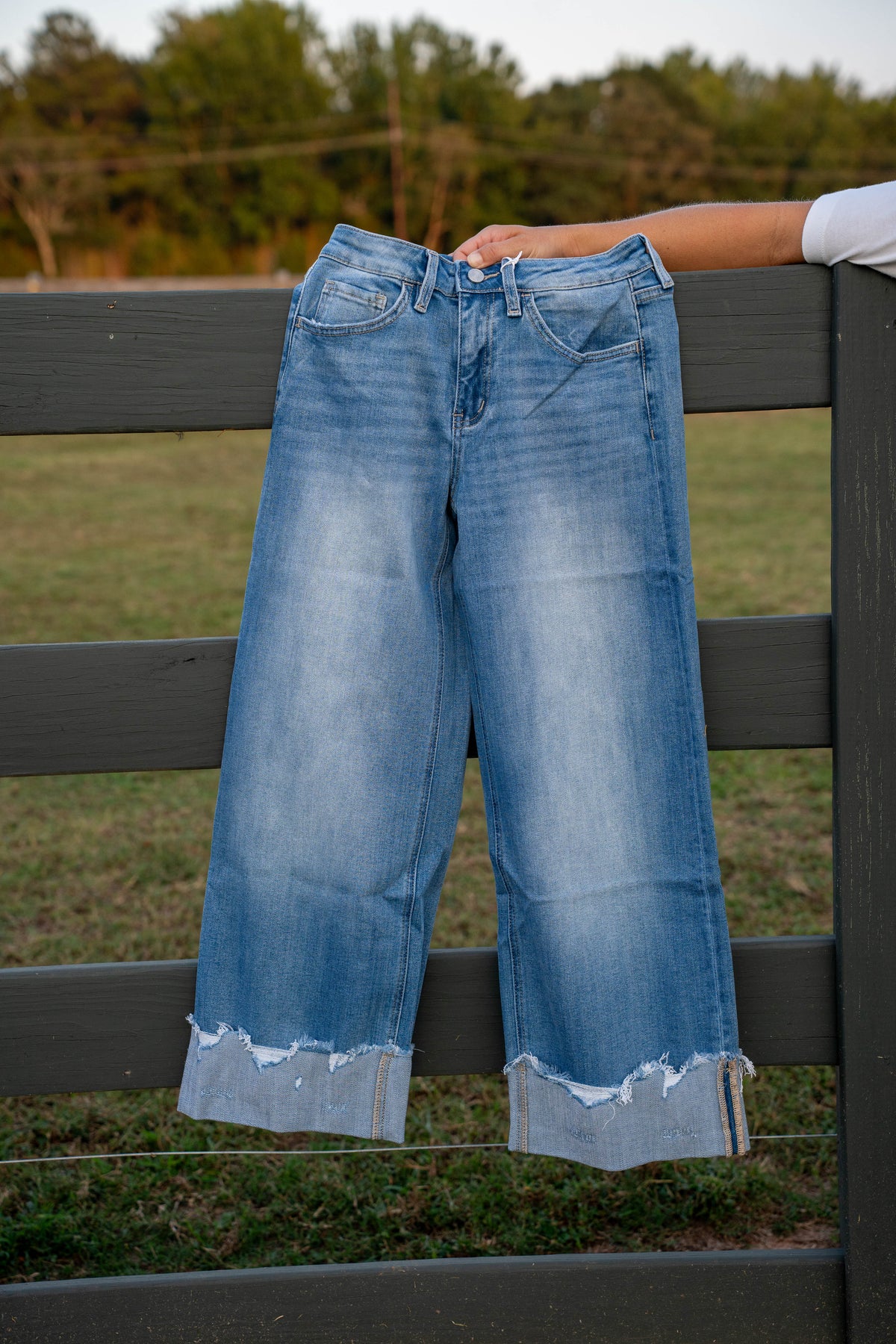 Light blue wide-leg cropped jeans with a raw, frayed hem, held up by a person's arm against a dark gray wooden fence in an outdoor grassy setting.
