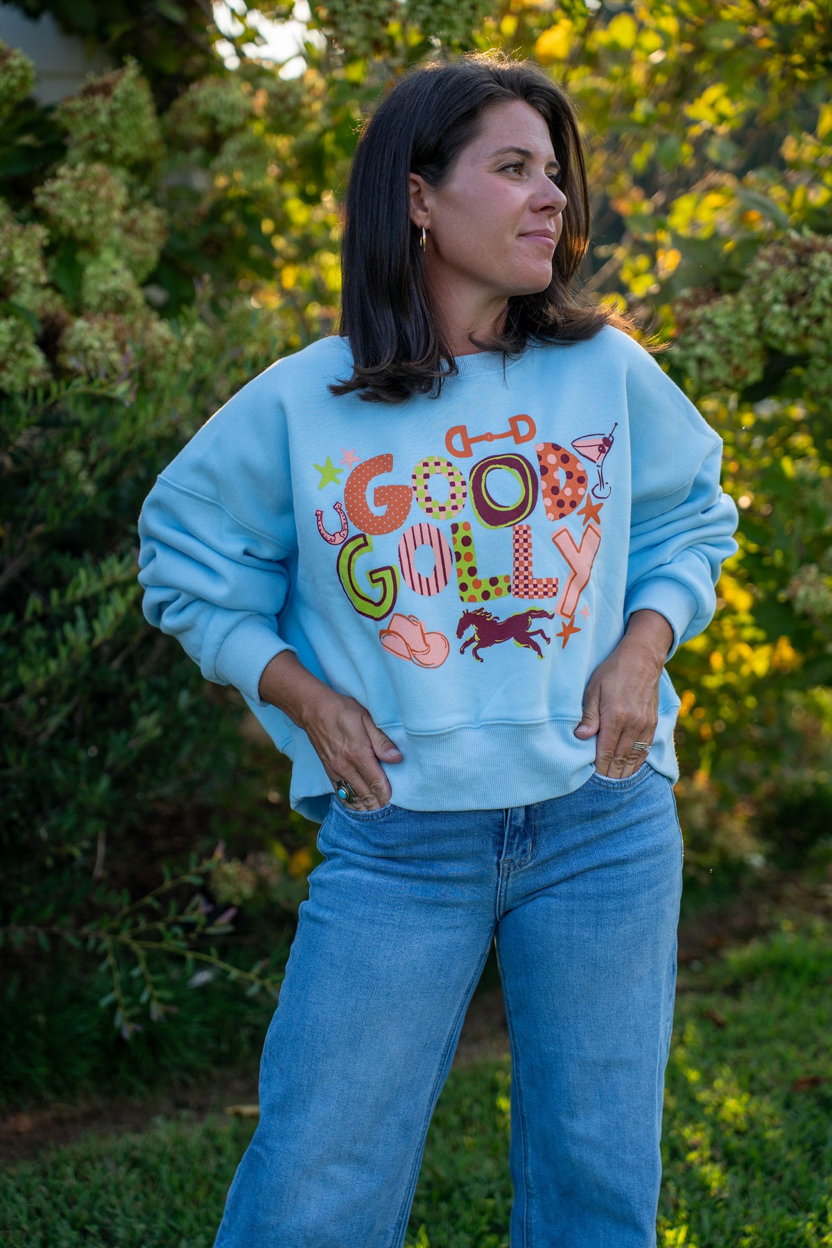 A woman wearing a light blue crewneck sweatshirt with a colorful retro 'Good Golly' graphic and blue jeans, standing outdoors in front of a blurred green garden background.