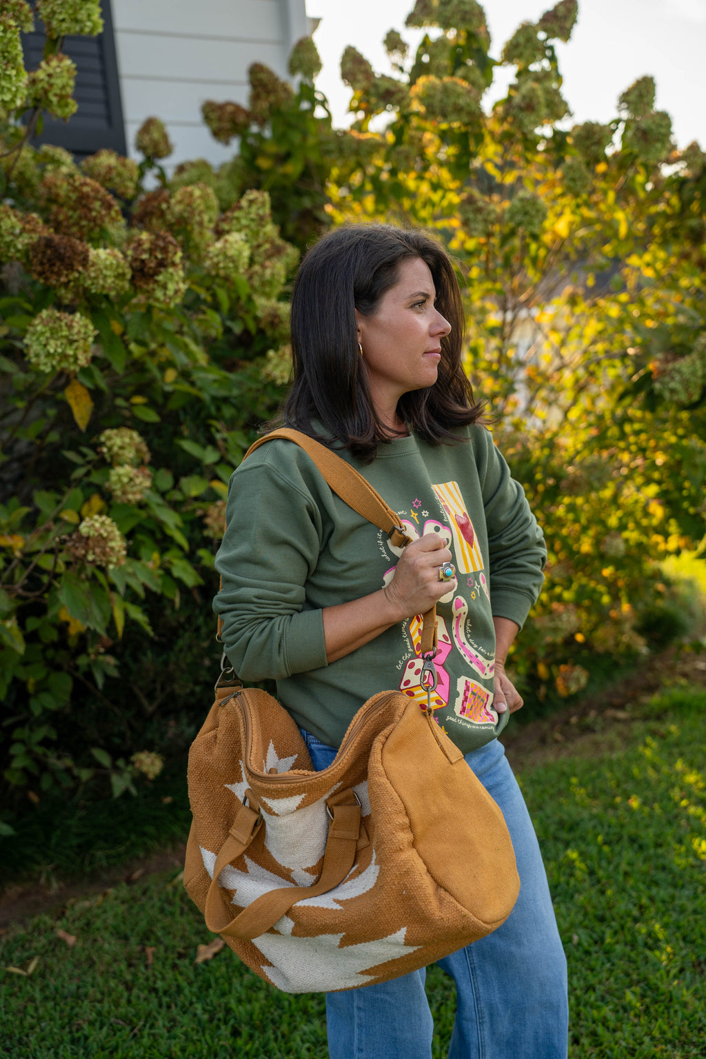  woman standing on a grassy lawn in front of leafy bushes, holding a tan Southwestern-style duffel bag with white geometric patterns. She is wearing a green graphic sweatshirt, blue jeans, and sandals.