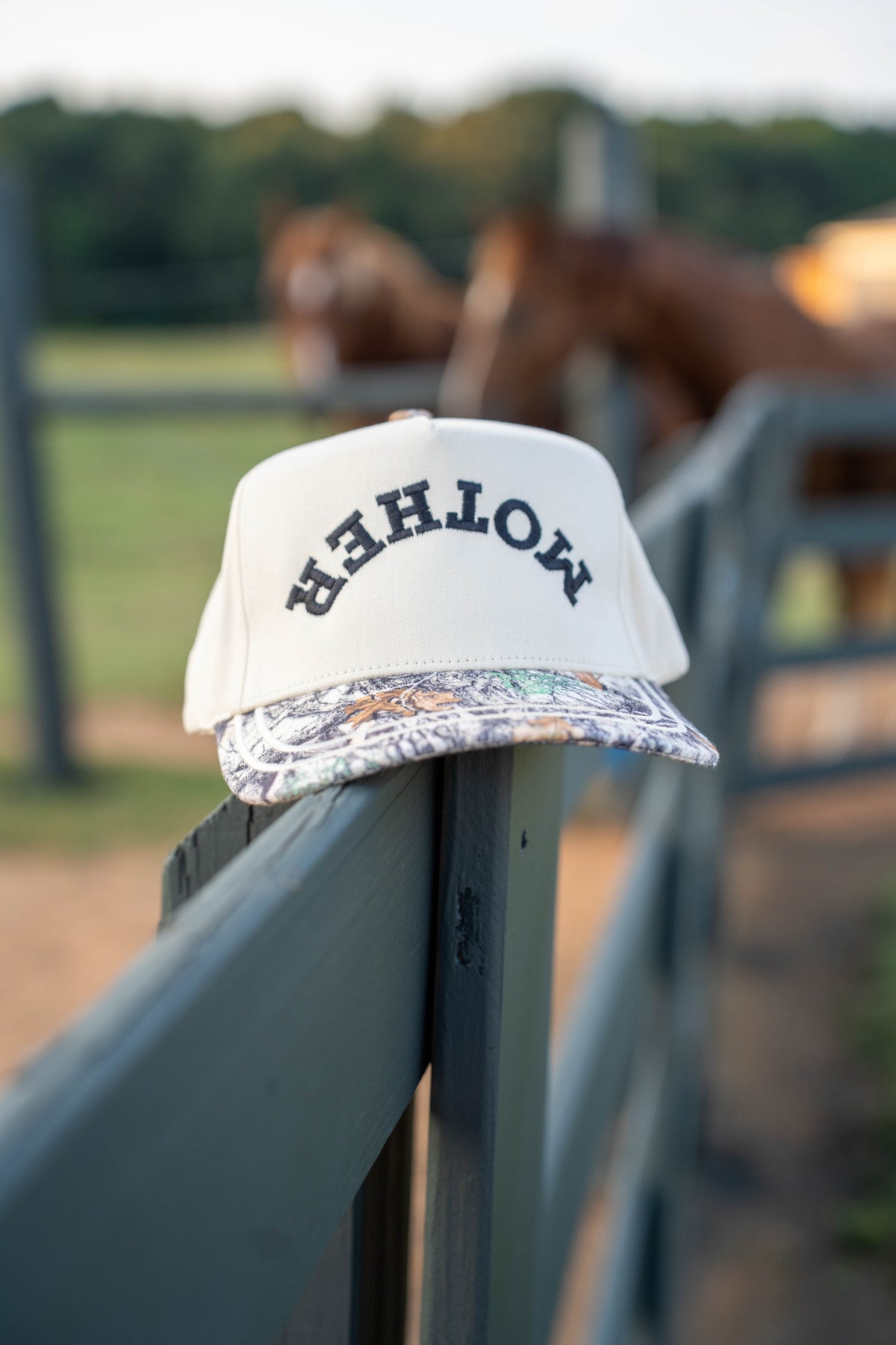 A white snapback cap with 'MOTHER' embroidered in black on the front, resting on a dark fence rail with a blurred outdoor ranch scene and horses in the background.
