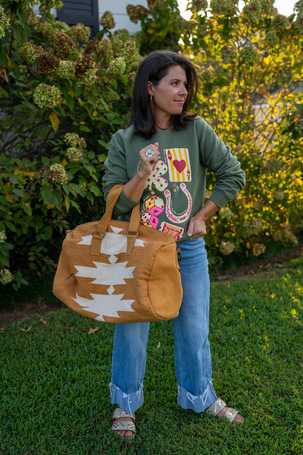A woman standing on a grassy lawn in front of leafy bushes, holding a tan Southwestern-style duffel bag with white geometric patterns. She is wearing a green graphic sweatshirt, blue jeans, and sandals.