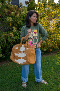 A woman standing on a grassy lawn in front of leafy bushes, holding a tan Southwestern-style duffel bag with white geometric patterns. She is wearing a green graphic sweatshirt, blue jeans, and sandals.