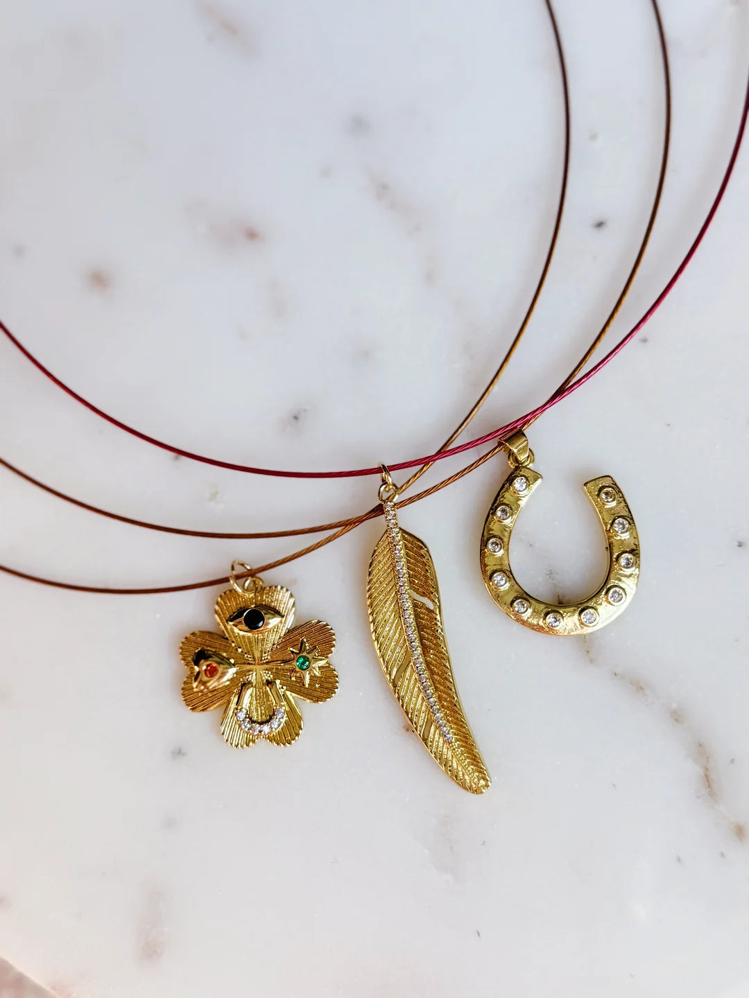 A close-up of a multi-strand necklace featuring three delicate gold-toned charms: a feather, a clover with small colored gem accents, and a horseshoe decorated with clear rhinestones, resting on a white marble surface.