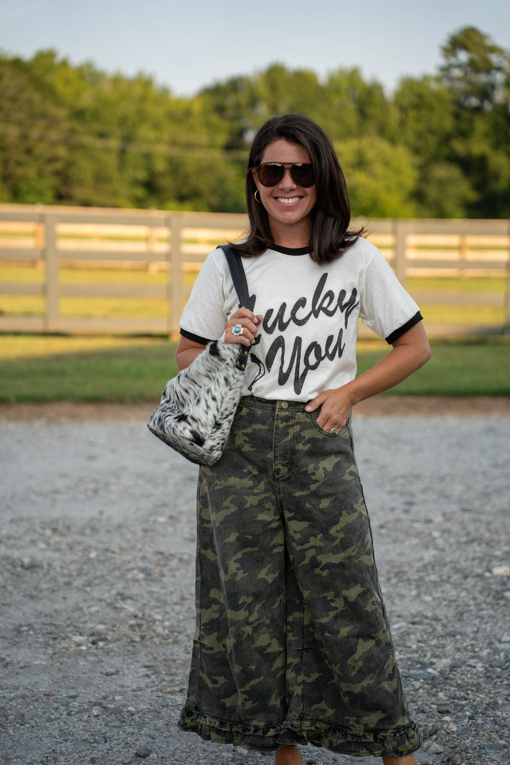 A woman stands outdoors on a gravel path wearing a white ringer T-shirt with black lettering that reads 'Lucky You,' paired with wide-leg dark green camouflage pants and carrying a black-and-white patterned shoulder bag.