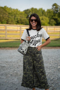 A woman stands outdoors on a gravel path wearing a white ringer T-shirt with black lettering that reads 'Lucky You,' paired with wide-leg dark green camouflage pants and carrying a black-and-white patterned shoulder bag.