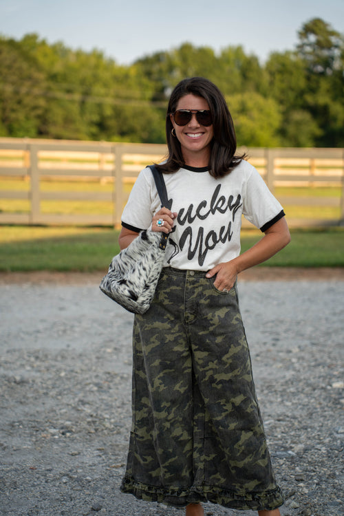 A woman stands outdoors on a gravel path wearing a white ringer T-shirt with black lettering that reads 'Lucky You,' paired with wide-leg dark green camouflage pants and carrying a black-and-white patterned shoulder bag.