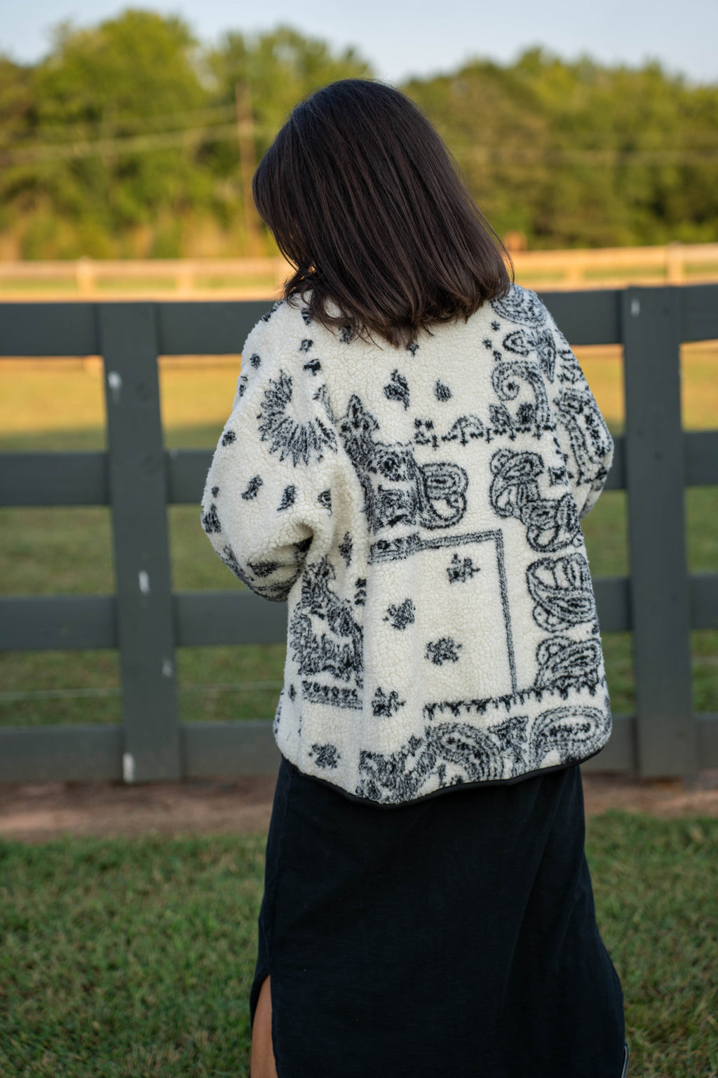 A woman viewed from behind wearing a cream-colored fuzzy cardigan with a black paisley and floral bandana-style print, standing outdoors in front of a dark gray wooden fence.