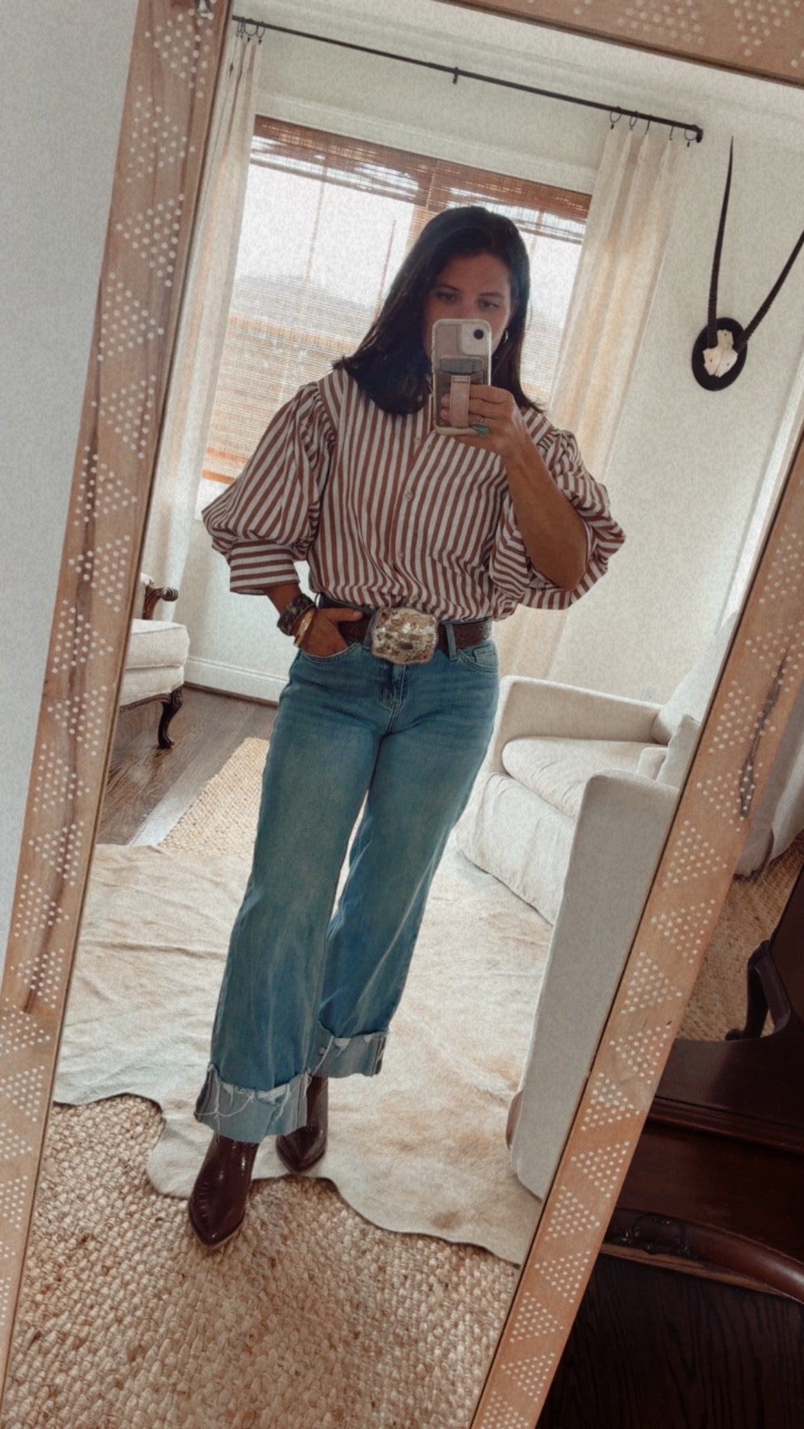 A woman takes a mirror selfie showing an outfit consisting of a brown and white vertically striped puff-sleeve blouse, light blue wide-leg jeans with rolled cuffs, a brown belt with a silver buckle, and brown boots, in a brightly lit room.