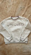 A light beige crewneck sweatshirt featuring the word 'UNIQUE' spelled out in horseshoe embroidery across the chest, laid flat on a brown faux fur surface.