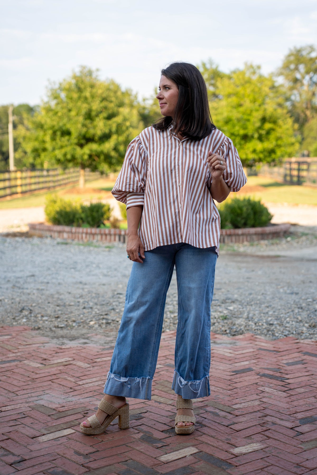 A woman stands outdoors on a brick path wearing a loose striped blouse with puff sleeves, blue wide-leg jeans with rolled cuffs, and tan heeled sandals. Trees and a fence are visible in the background.