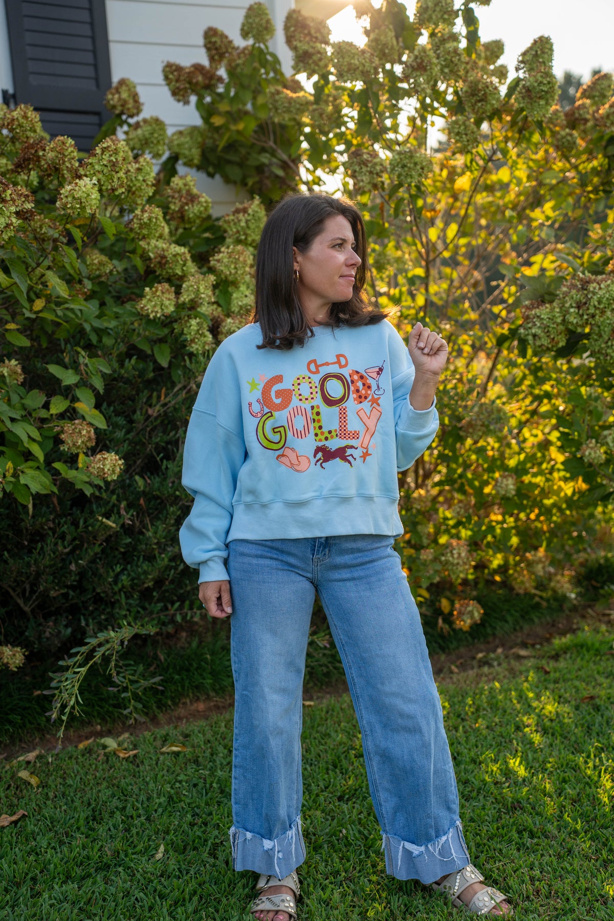 A woman stands outdoors on a grassy lawn in front of leafy bushes, wearing a light blue sweatshirt with a colorful retro-style graphic that reads 'GOD GULY' and light-wash wide-leg jeans.