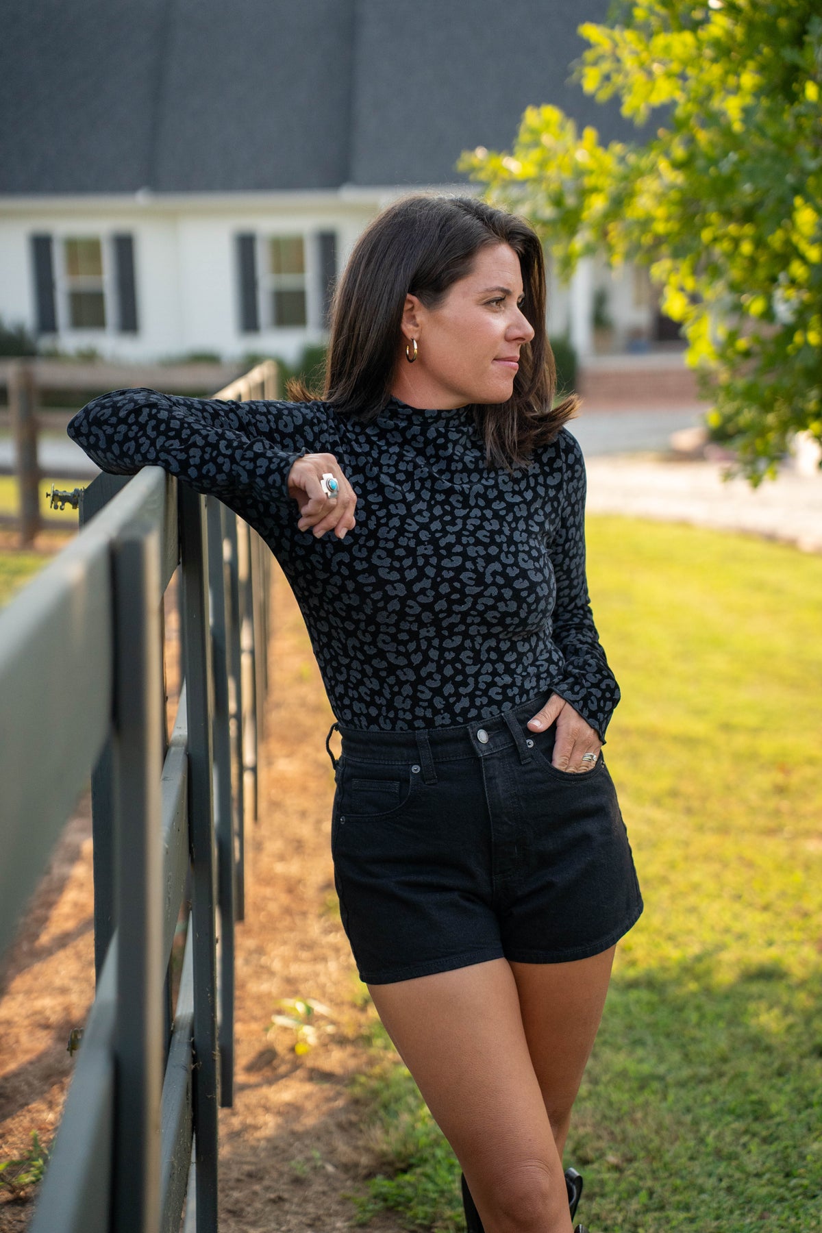 A woman wearing a black long-sleeve top with a small gray leopard print and black denim shorts, leaning against a wooden fence in a backyard setting.
