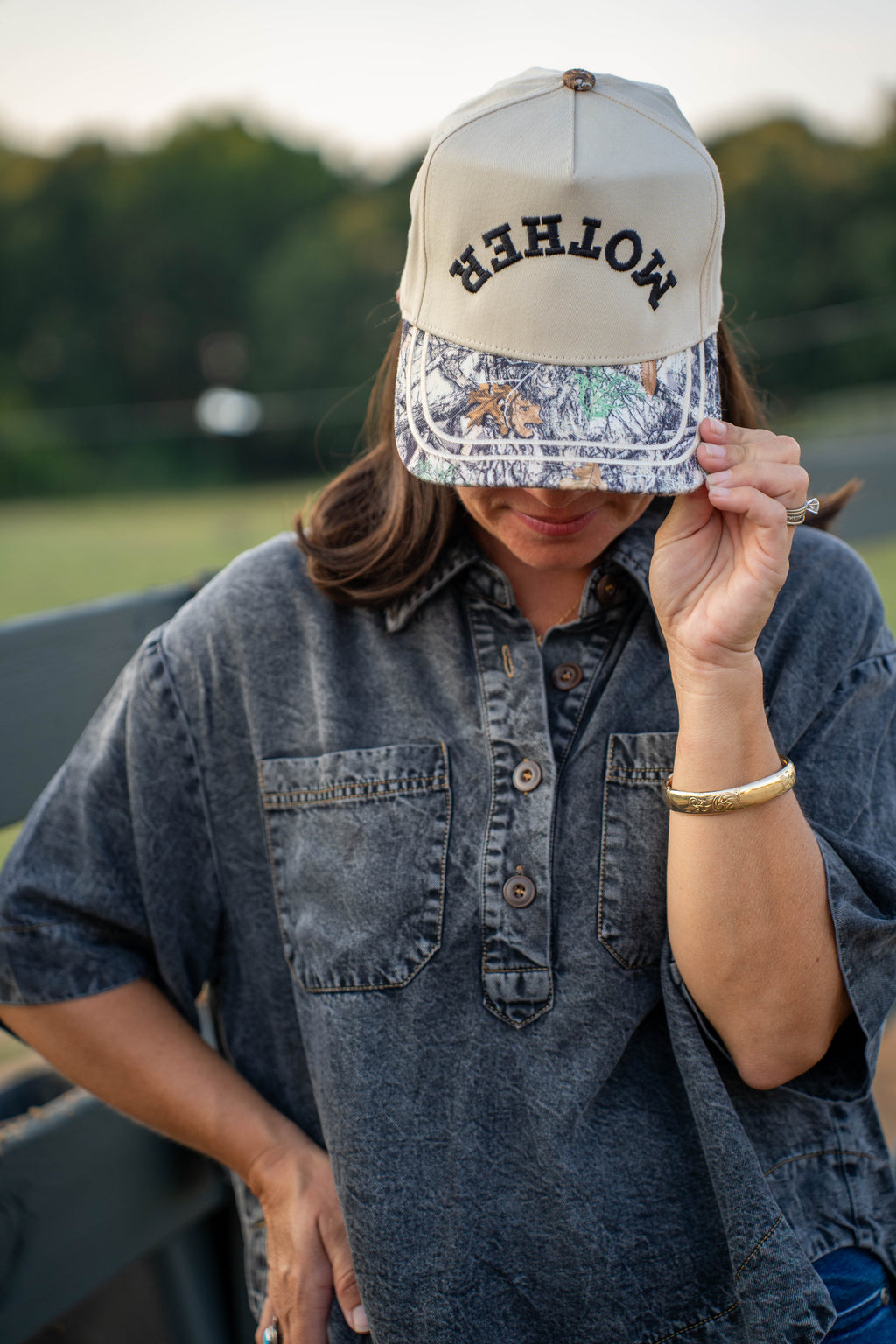 A white snapback cap with 'MOTHER' embroidered in black on the front, resting on a dark fence rail with a blurred outdoor ranch scene and horses in the background.
