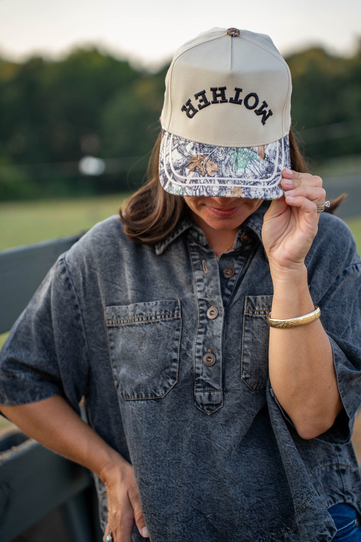 A white snapback cap with 'MOTHER' embroidered in black on the front, resting on a dark fence rail with a blurred outdoor ranch scene and horses in the background.