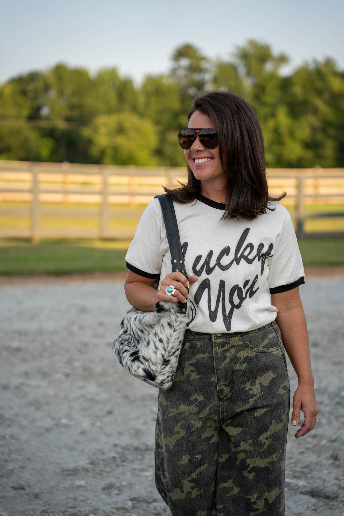 A woman wearing a white ringer t-shirt with black lettering that reads 'Lucky You', paired with camouflage pants and sunglasses, carrying a black and white patterned shoulder bag in an outdoor setting.