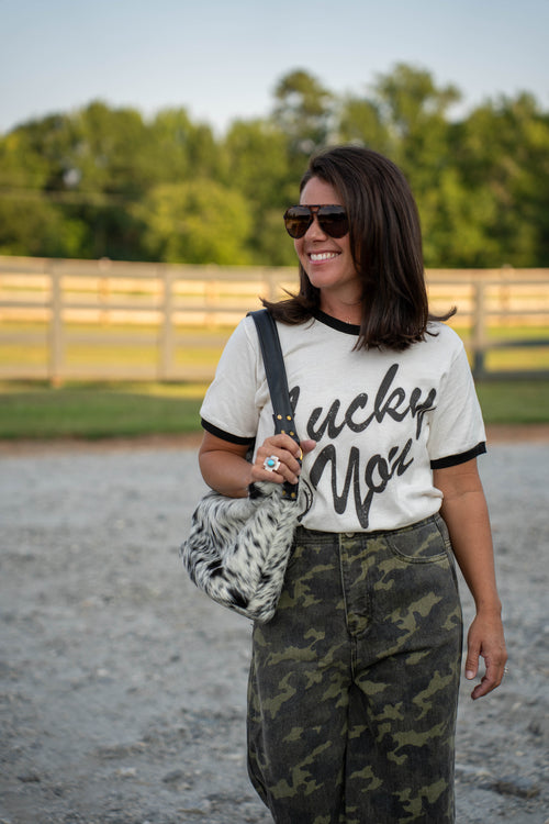 A woman wearing a white ringer t-shirt with black lettering that reads 'Lucky You', paired with camouflage pants and sunglasses, carrying a black and white patterned shoulder bag in an outdoor setting.