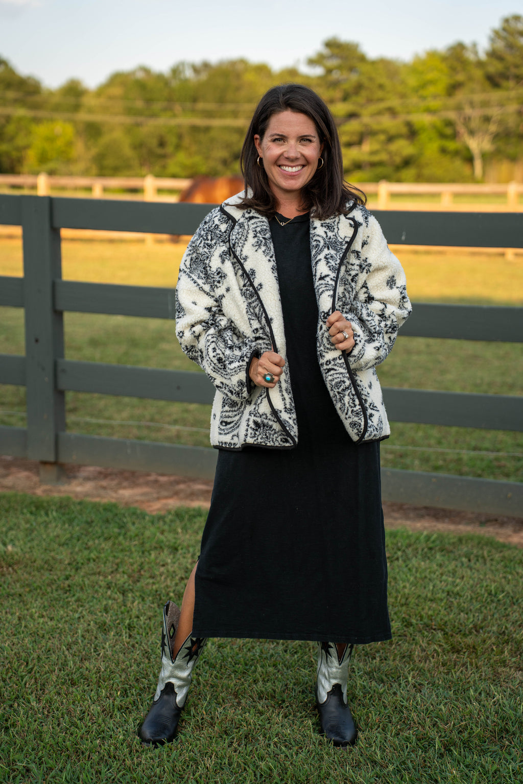 A woman stands outdoors on grass in front of a wooden fence, wearing a black dress with a black-and-white patterned jacket, black boots, and light-colored cowboy boots.