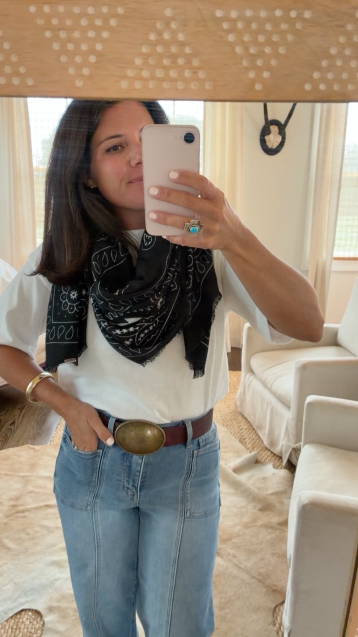 A woman takes a mirror selfie showing a styled outfit with a black and white bandana scarf, a white t-shirt, light blue jeans with a brown belt, and a large gold buckle. She is wearing a turquoise ring and gold bracelets.