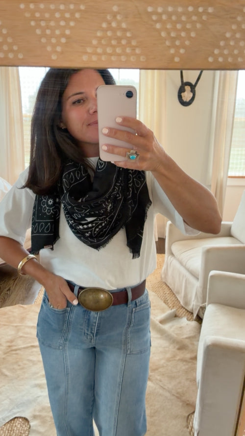 A woman takes a mirror selfie showing a styled outfit with a black and white bandana scarf, a white t-shirt, light blue jeans with a brown belt, and a large gold buckle. She is wearing a turquoise ring and gold bracelets.